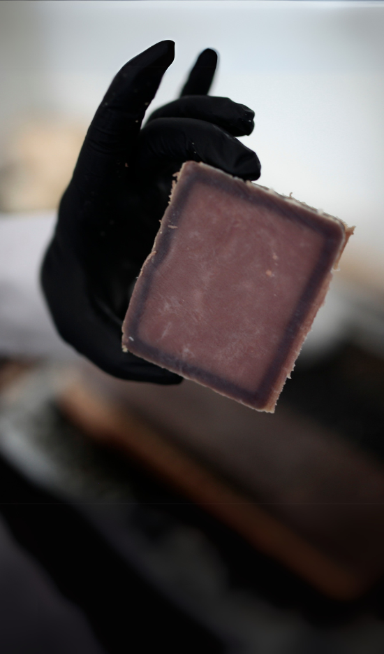 Hand in schwarzem Latexhandschuh hält eine frische, dunkle Naturseife hoch