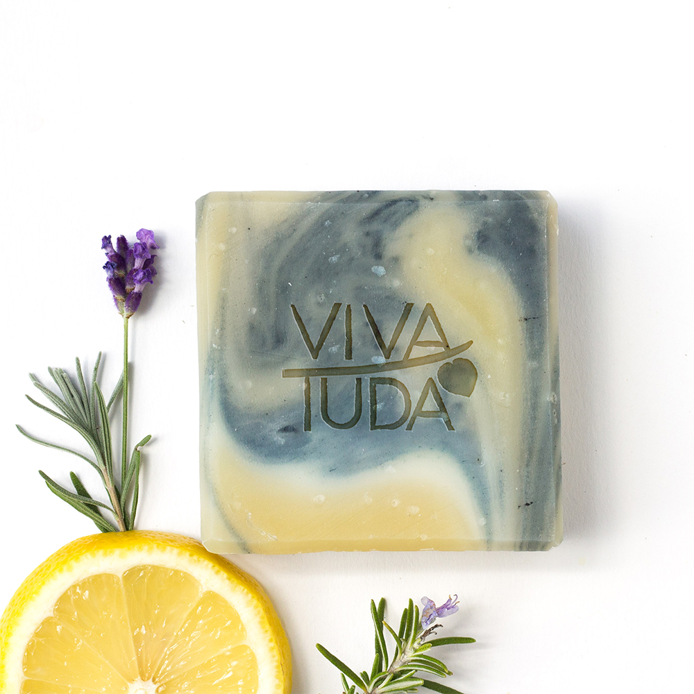 Hellgelb und blau-grau marmorierte Naturseife, daneben eine Zitronenscheibe und Rosmarin mit Blüte.