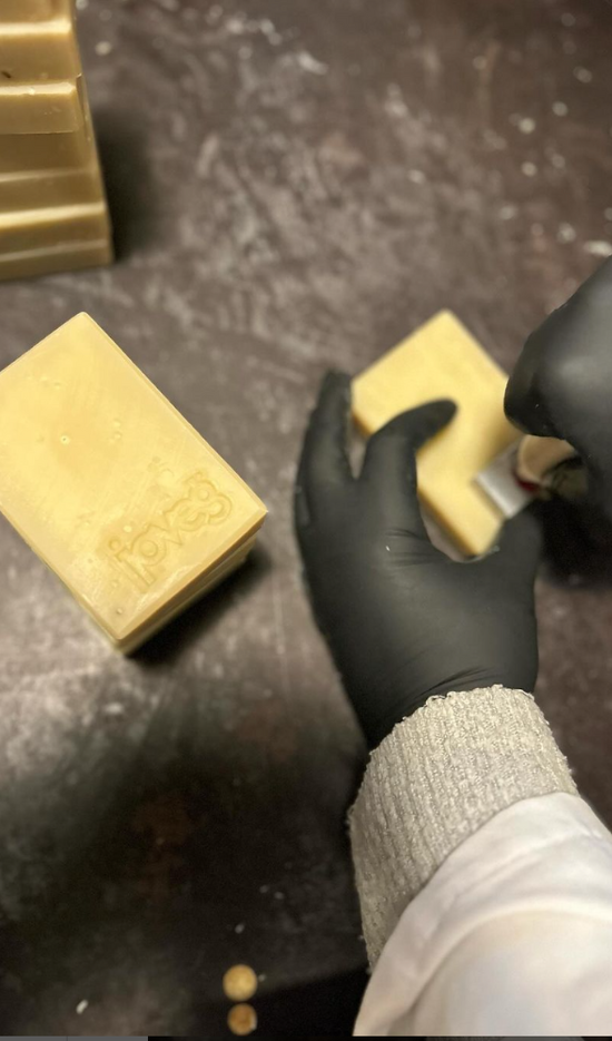 Hände in schwarzen Latexhandschuhen stempeln eine gelbliche Seife. Im Hintergrund eine gestempelte Seifen und ein Stapel weiterer Seifen