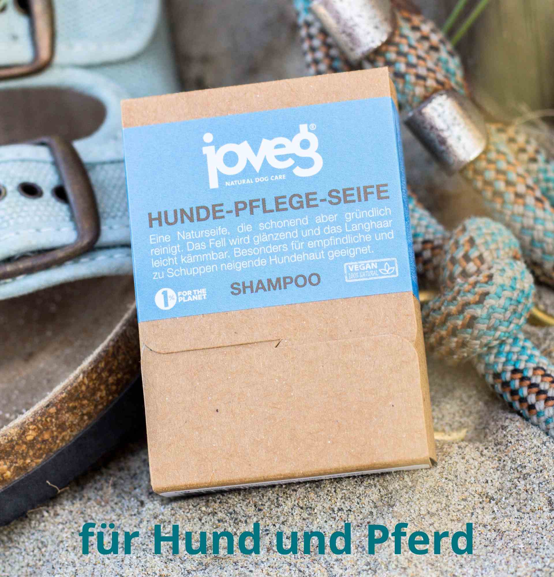 Hundeshampooseife joveg, mit blauem Etikett im Sand, Sandalen und Hundeleine im Hintergrund