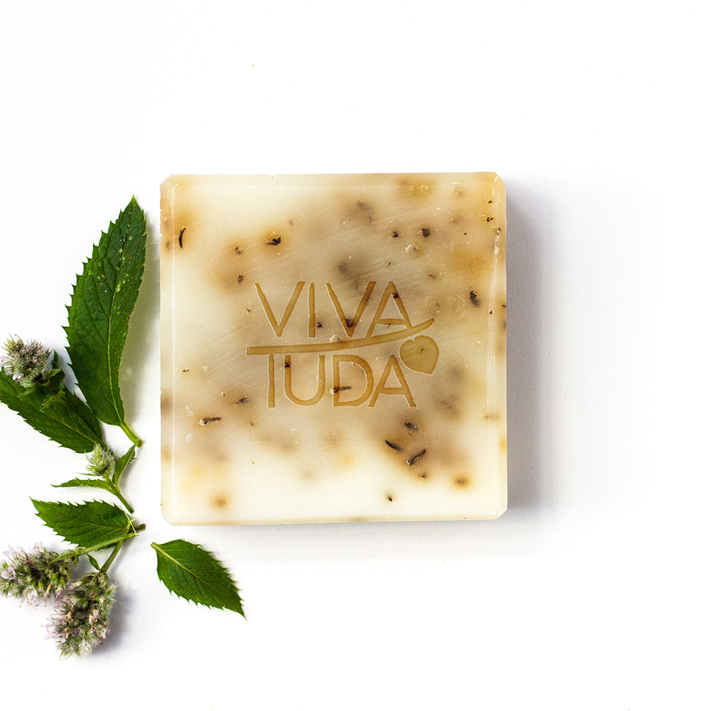 Helle Naturseife mit hellbrauner Marmorierung und Kräutern, daneben Minzblätter und Blüten.